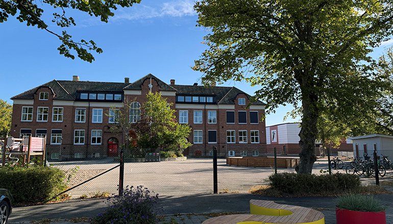 Stor rödbrun skolbyggnad, staket framför.